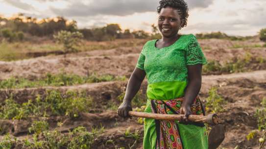 A woman (Faith Muvili) stands in a farm field holding a hoe, looking at the camera.