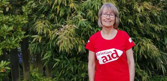 Image of a white woman, stood against a green garden background, wearing a red branded Christian Aid t-shirt