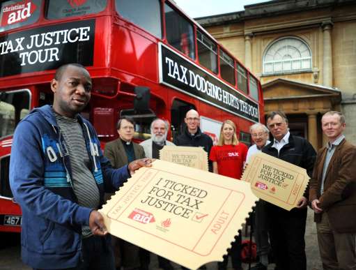 Savior Mwamba from Zambia holding a ticket saying "I ticked for tax justice" alongside a group of other campaigners in front of a red bus with the words "Tax justice tour" displayed on the front.