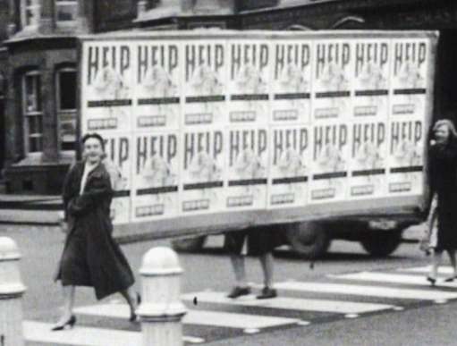 Women cross the road carrying a large banner covered in posters with 'help' written on them.