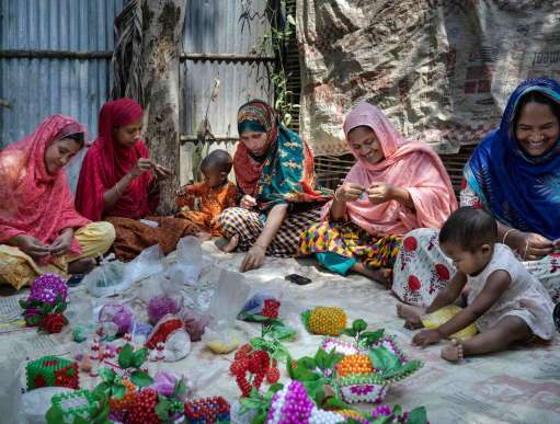 A group of 5 women in colourful garments in Bangladesh sit on the floor working with beads. Two small babies sit with them playing. 