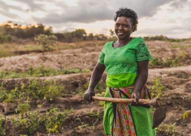 A woman (Faith Muvili) stands in a farm field holding a hoe, looking at the camera.