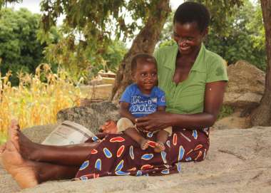 A smiling young black woman, wearing a green top, patterned skirt and sat barefoot on the ground, holds her smiling baby son